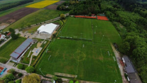 Alttext: Luftaufnahme der Hans-Dorfner-Fußballschule mit mehreren Soccerfeldern, Trainingsplätzen und Vereinsgebäuden in einer ländlichen Umgebung, umgeben von Wäldern und Feldern.
