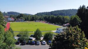 Fußballschule Hans Dorfner auf einem grünen Sportplatz, umgeben von Bäumen und Bergen, ideale Trainingsbedingungen für Kinder und Jugendliche im Fußball, inklusive Laufbahn und schöner Naturkulisse.