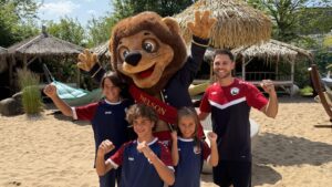 Fröhliche Kinder und Trainer mit Maskottchen auf einem sandigen Fußballplatz.